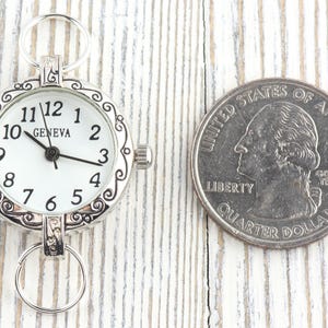 Pode incluir: Um rel&oacute;gio de bolso Geneva prateado com mostrador branco e algarismos pretos, ao lado de uma moeda de um quarto de d&oacute;lar americano. O rel&oacute;gio tem uma moldura decorativa e dois an&eacute;is para fixa&ccedil;&atilde;o. A moeda mostra George Washington.