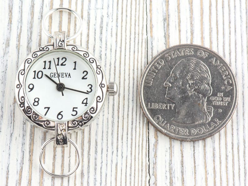 Pode incluir: Um rel&oacute;gio de bolso Geneva prateado com mostrador branco e algarismos pretos, ao lado de uma moeda de um quarto de d&oacute;lar americano. O rel&oacute;gio tem uma moldura decorativa e dois an&eacute;is para fixa&ccedil;&atilde;o. A moeda mostra George Washington.