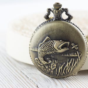 Pode incluir: Um rel&oacute;gio de bolso dourado com um design detalhado de um peixe saltando para fora da &aacute;gua. O rel&oacute;gio tem uma corrente decorativa.