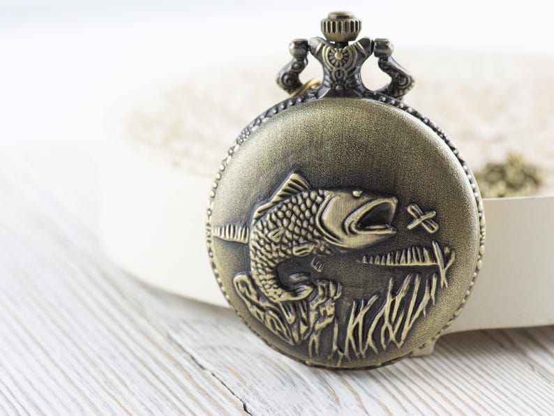 Pode incluir: Um rel&oacute;gio de bolso dourado com um design detalhado de um peixe saltando para fora da &aacute;gua. O rel&oacute;gio tem uma corrente decorativa.