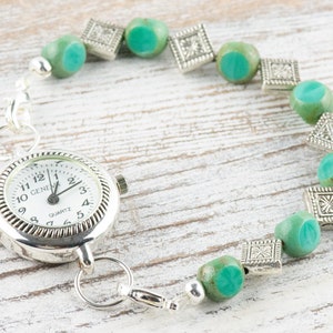 Pu&ograve; includere: Un orologio da tasca argentato con quadrante bianco e numeri neri &egrave; attaccato a un braccialetto fatto di perline blu turchese e distanziatori argentati.