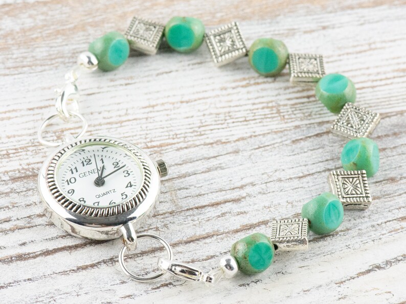 Pu&ograve; includere: Un orologio da tasca argentato con quadrante bianco e numeri neri &egrave; attaccato a un braccialetto fatto di perline blu turchese e distanziatori argentati.