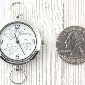Puede incluir: Un reloj de bolsillo plateado Geneva con esfera blanca, contornos de mariposas negras y los n&uacute;meros 12, 3, 6 y 9. El reloj tiene un anillo met&aacute;lico circular en la parte superior e inferior, colocado junto a una moneda de cuarto de d&oacute;lar.