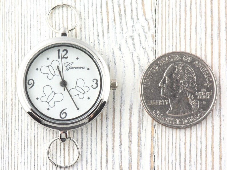 Puede incluir: Un reloj de bolsillo plateado Geneva con esfera blanca, contornos de mariposas negras y los n&uacute;meros 12, 3, 6 y 9. El reloj tiene un anillo met&aacute;lico circular en la parte superior e inferior, colocado junto a una moneda de cuarto de d&oacute;lar.