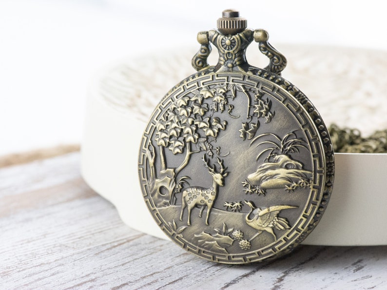 Pu&ograve; includere: Un orologio da tasca in bronzo con un design elaborato che presenta un cervo, un uccello e un albero. L'orologio ha una catena.