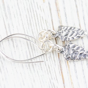 K&ouml;nnte beinhalten: Silberne Ohrringe mit einem strukturierten Pfeilspitzen-Design. Die Ohrringe sind an einer silbernen Kette befestigt und haben einen einfachen Hakenverschluss.