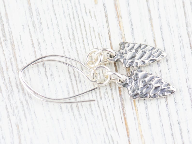 K&ouml;nnte beinhalten: Silberne Ohrringe mit einem strukturierten Pfeilspitzen-Design. Die Ohrringe sind an einer silbernen Kette befestigt und haben einen einfachen Hakenverschluss.