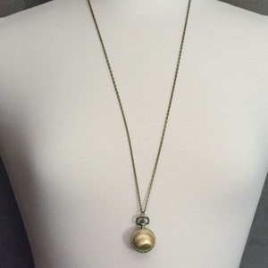 May include: A gold-toned pocket watch pendant on a chain necklace. The watch has a round face and a small, decorative knob.