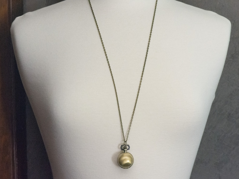 May include: A gold-toned pocket watch pendant on a chain necklace. The watch has a round face and a small, decorative knob.