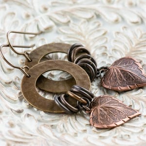 Op de afbeelding: Handgemaakte oorbellen met bronzen cirkelvormige ringen en bladhangers. De oorbellen hebben een rustieke, vintage stijl, met gedetailleerde bladbedels. De sieraden liggen op een witte, getextureerde ondergrond.