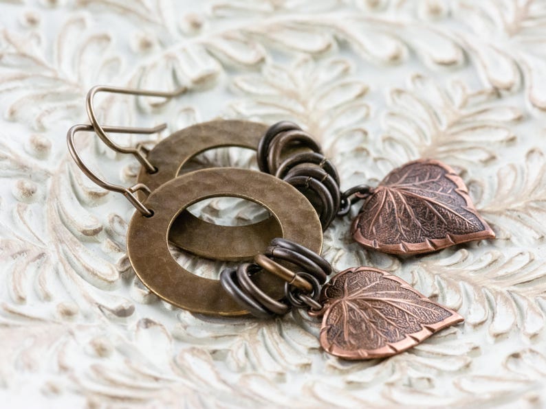 Op de afbeelding: Handgemaakte oorbellen met bronzen cirkelvormige ringen en bladhangers. De oorbellen hebben een rustieke, vintage stijl, met gedetailleerde bladbedels. De sieraden liggen op een witte, getextureerde ondergrond.