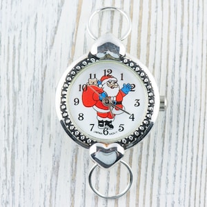Peut inclure: Une montre de poche argent&eacute;e avec un cadran blanc repr&eacute;sentant un P&egrave;re No&euml;l dessin anim&eacute; tenant un sac de cadeaux. La montre a une boucle en forme de c&oelig;ur et une cha&icirc;ne argent&eacute;e.
