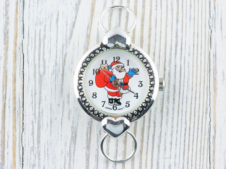 Peut inclure: Une montre de poche argent&eacute;e avec un cadran blanc repr&eacute;sentant un P&egrave;re No&euml;l dessin anim&eacute; tenant un sac de cadeaux. La montre a une boucle en forme de c&oelig;ur et une cha&icirc;ne argent&eacute;e.