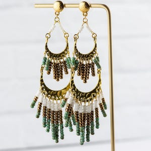 May include: Gold hoop earrings with a layered design featuring white, brown, and green beads. The earrings have a bohemian style.
