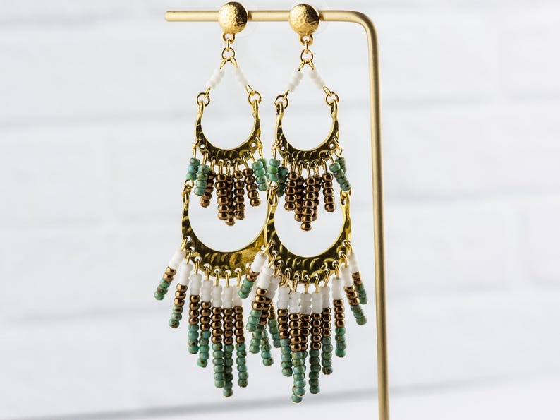 May include: Gold hoop earrings with a layered design featuring white, brown, and green beads. The earrings have a bohemian style.