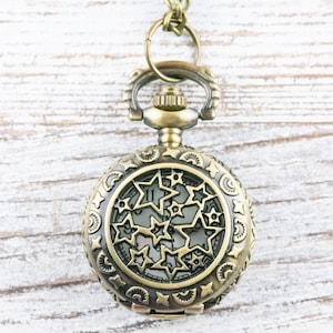 May include: A gold-toned pocket watch with a star-shaped design on the face. The watch is hanging from a chain on a white wooden background.