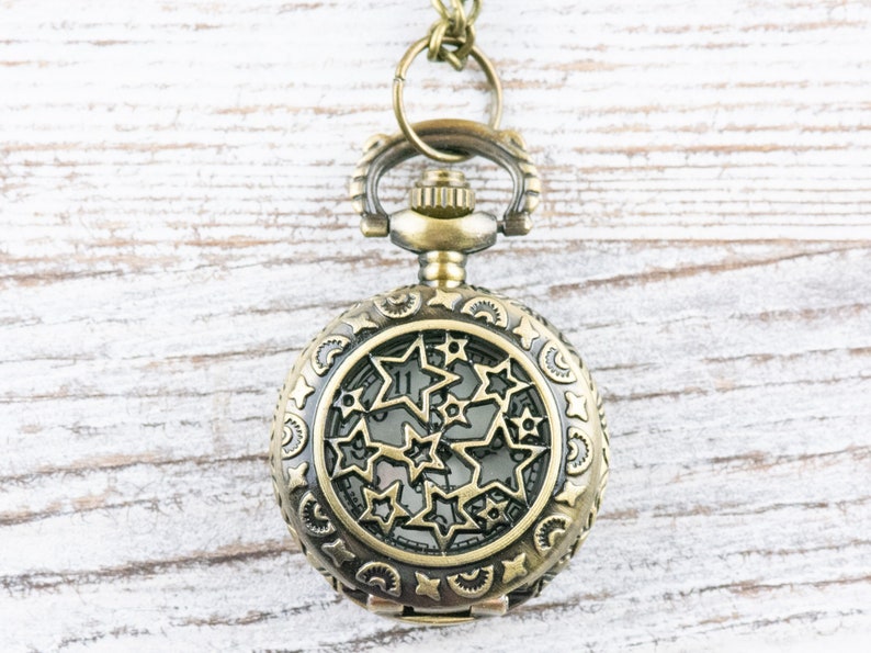 May include: A gold-toned pocket watch with a star-shaped design on the face. The watch is hanging from a chain on a white wooden background.