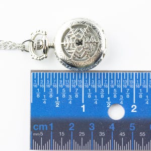 May include: A silver pocket watch with a snowflake design on the face. The watch is hanging from a silver chain and is measured against a ruler.
