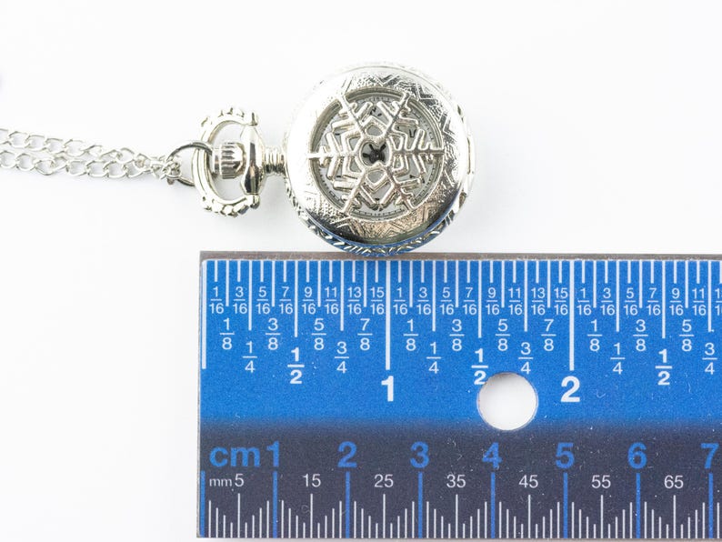May include: A silver pocket watch with a snowflake design on the face. The watch is hanging from a silver chain and is measured against a ruler.