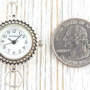K&ouml;nnte beinhalten: Eine kleine, silberfarbene Taschenuhr mit wei&szlig;em Zifferblatt und schwarzen Zeigern, neben einem US-Vierteldollar. Die Uhr hat eine dekorative L&uuml;nette und zwei kleine Ringe zur Befestigung. Die M&uuml;nze dient zum Gr&ouml;&szlig;envergleich.