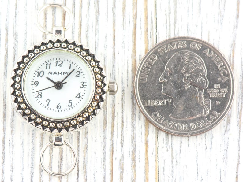 K&ouml;nnte beinhalten: Eine kleine, silberfarbene Taschenuhr mit wei&szlig;em Zifferblatt und schwarzen Zeigern, neben einem US-Vierteldollar. Die Uhr hat eine dekorative L&uuml;nette und zwei kleine Ringe zur Befestigung. Die M&uuml;nze dient zum Gr&ouml;&szlig;envergleich.