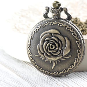 May include: Antique bronze pocket watch with a rose design on the cover. The watch has a decorative border and a chain. The watch is a vintage style accessory.