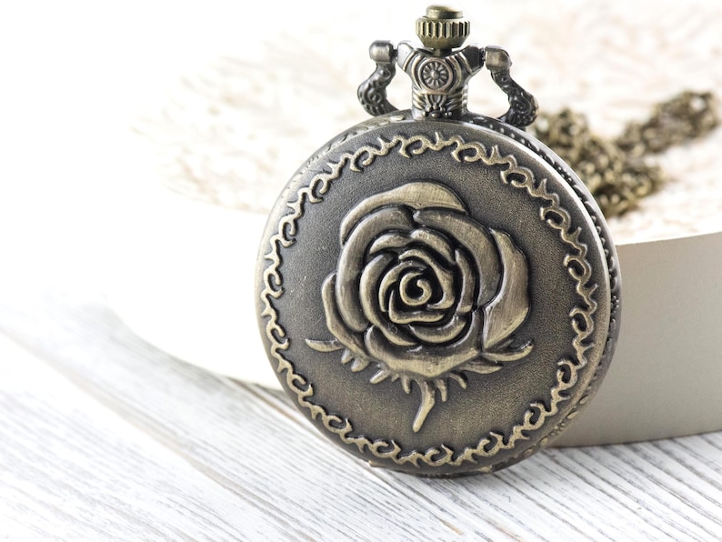 May include: Antique bronze pocket watch with a rose design on the cover. The watch has a decorative border and a chain. The watch is a vintage style accessory.