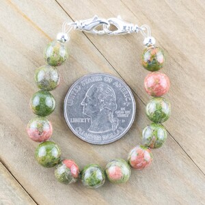 Puede incluir: Un brazalete de piedras preciosas verdes y rosas con un cierre plateado. El brazalete est&aacute; hecho de cuentas redondas y tiene aproximadamente el tama&ntilde;o de una moneda de 25 c&eacute;ntimos de euro.