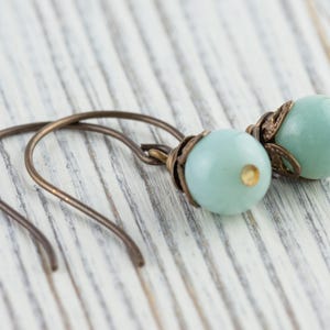 May include: A pair of earrings featuring pale blue, round beads with bronze-colored metal accents. The earrings have a simple hook design and are displayed on a white wooden surface.