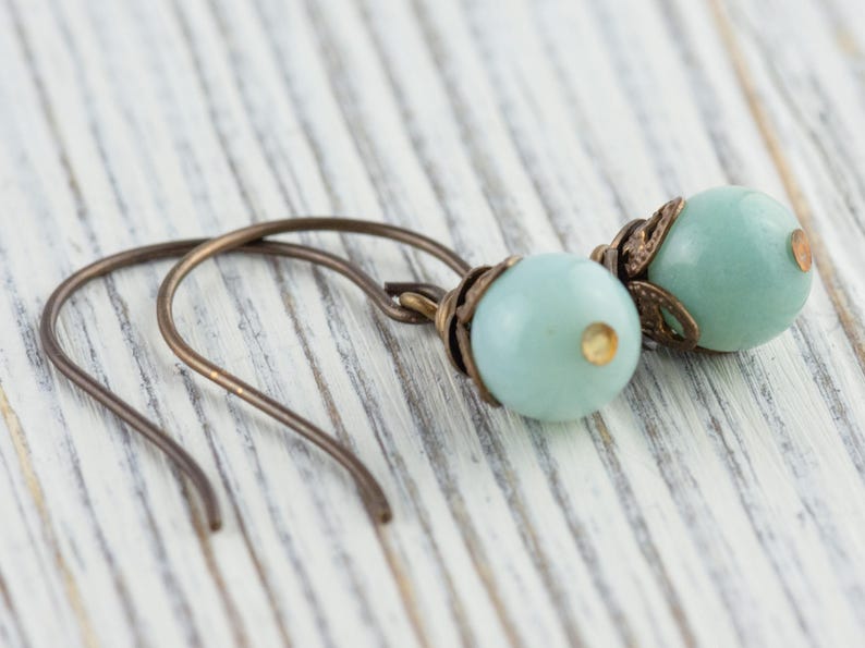 May include: A pair of earrings featuring pale blue, round beads with bronze-colored metal accents. The earrings have a simple hook design and are displayed on a white wooden surface.