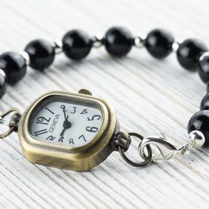 Pu&ograve; includere: Un braccialetto di perline nere con un orologio dorato attaccato. Il quadrante dell'orologio &egrave; quadrato e ha le marcature dei numeri romani. Il quadrante dell'orologio ha anche la parola "GENEVA" stampata.