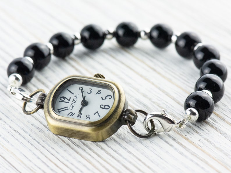 Pu&ograve; includere: Un braccialetto di perline nere con un orologio dorato attaccato. Il quadrante dell'orologio &egrave; quadrato e ha le marcature dei numeri romani. Il quadrante dell'orologio ha anche la parola "GENEVA" stampata.
