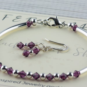 May include: A silver bangle bracelet with a row of purple crystal beads and a matching pair of dangle earrings with purple crystal beads. The bracelet has a silver clasp.