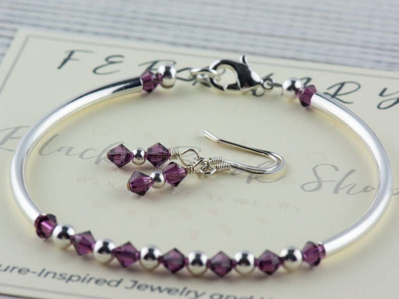 May include: A silver bangle bracelet with a row of purple crystal beads and a matching pair of dangle earrings with purple crystal beads. The bracelet has a silver clasp.