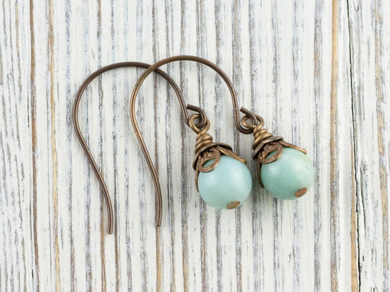 May include: A pair of earrings with a bronze-colored hook and decorative top. Each earring features a light blue, round bead. The earrings are displayed on a white, weathered wooden surface.