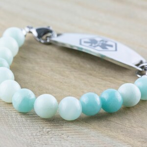 May include: A medical ID bracelet with a silver tag and a blue and white beaded bracelet. The tag has a medical symbol on it.