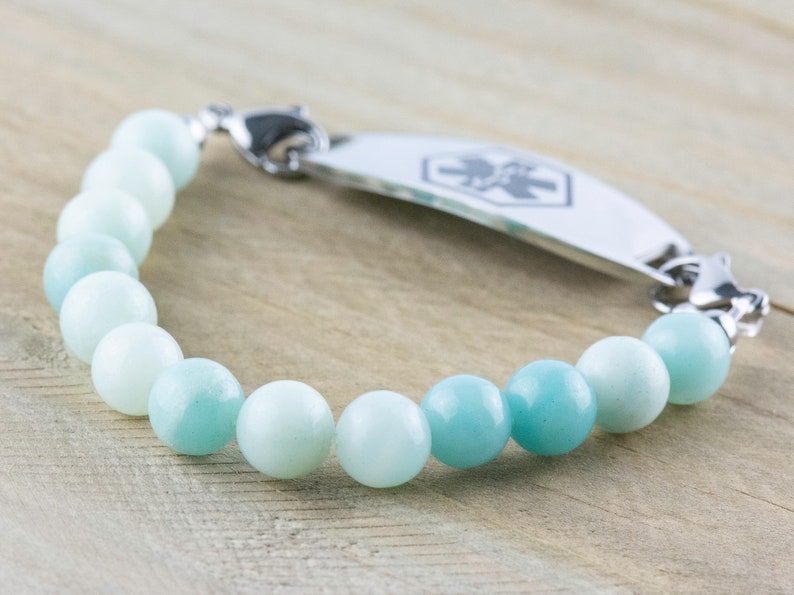 May include: A medical ID bracelet with a silver tag and a blue and white beaded bracelet. The tag has a medical symbol on it.