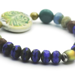 Puede incluir: Un brazalete de cuentas azules y verdes con un dije de cer&aacute;mica con un dise&ntilde;o de hojas.