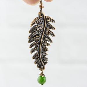 K&ouml;nnte beinhalten: Ein Paar bronzefarbene Ohrringe in Form eines Farnblattes mit einem gr&uuml;nen Jade-Tropfen. Die Ohrringe sind aus Metall und haben ein detailliertes Farnblatt-Design.