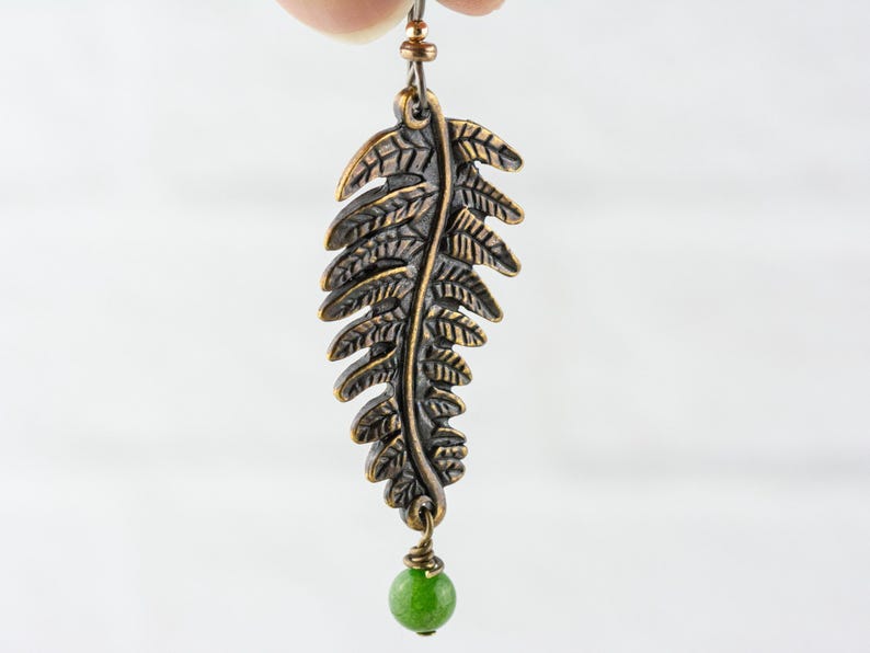 K&ouml;nnte beinhalten: Ein Paar bronzefarbene Ohrringe in Form eines Farnblattes mit einem gr&uuml;nen Jade-Tropfen. Die Ohrringe sind aus Metall und haben ein detailliertes Farnblatt-Design.