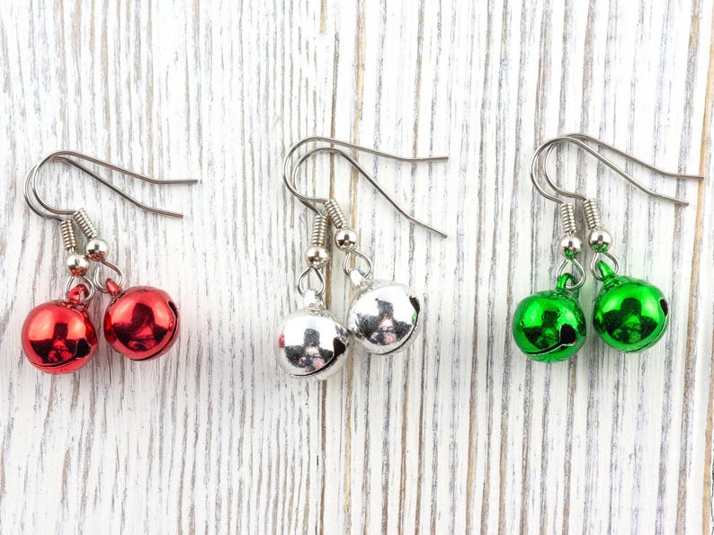 May include: Three pairs of bell earrings in red, silver, and green. Each pair features a small, shiny bell suspended from a silver hook. The earrings are displayed on a white, weathered wooden surface.