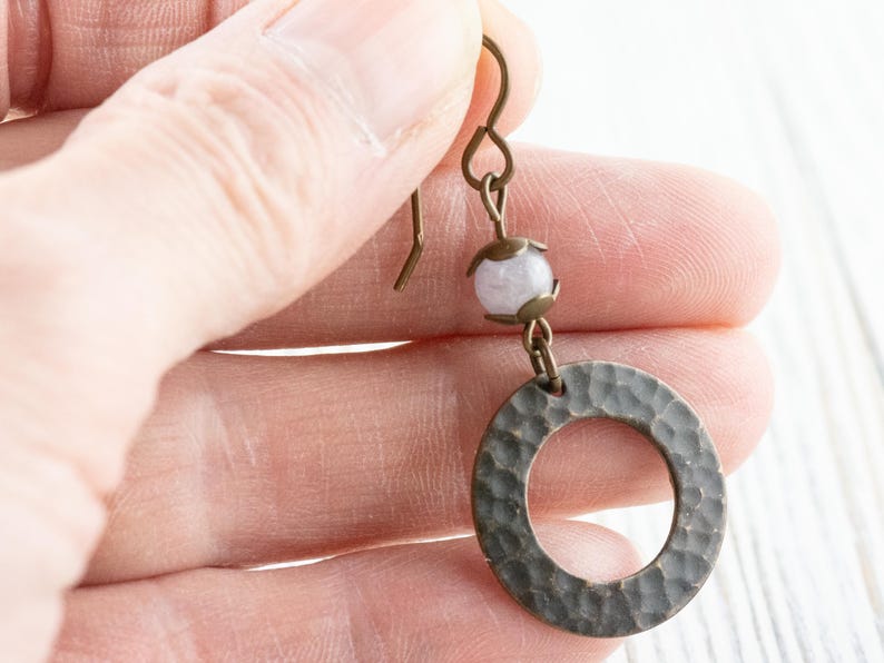 Puede incluir: Un solo pendiente con un c&iacute;rculo de metal martillado y una peque&ntilde;a cuenta de color claro. El pendiente tiene un gancho y elementos de conexi&oacute;n de color bronce. El c&iacute;rculo tiene una superficie texturizada.