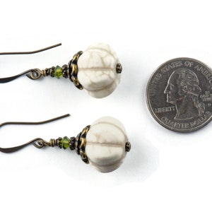 May include: A pair of white, textured, round earrings with green glass beads and bronze accents. The earrings are shown next to a UK 20 pence coin for size comparison.