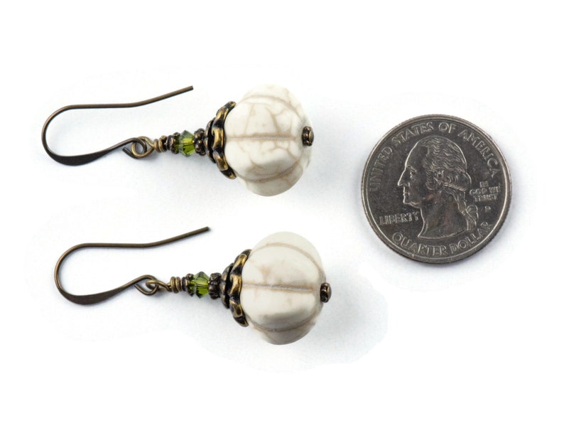 May include: A pair of white, textured, round earrings with green glass beads and bronze accents. The earrings are shown next to a UK 20 pence coin for size comparison.