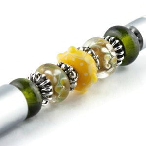 Peut inclure: Un stylo argent&eacute; avec un charme de perles en verre vert, clair et jaune. Les perles ont un motif floral et des pois blancs.