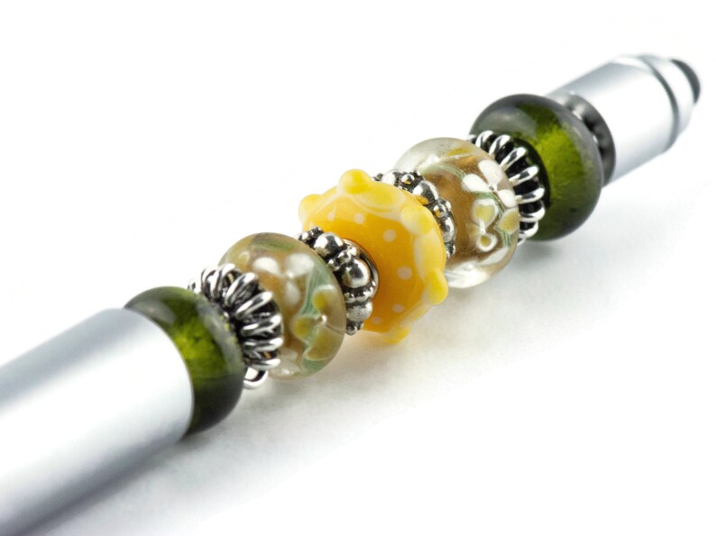 Peut inclure: Un stylo argent&eacute; avec un charme de perles en verre vert, clair et jaune. Les perles ont un motif floral et des pois blancs.