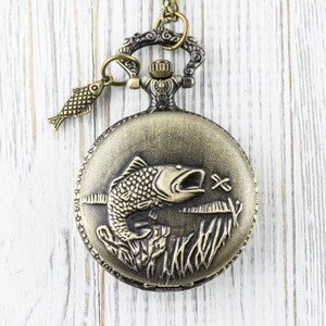 Pode incluir: Um rel&oacute;gio de bolso dourado com uma corrente e um pequeno amuleto de peixe. O mostrador do rel&oacute;gio apresenta uma imagem gravada de um peixe saltando para fora da &aacute;gua.