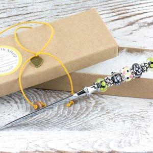 K&ouml;nnte beinhalten: Ein Brief&ouml;ffner mit einem dekorativen, mit Perlen besetzten Griff, pr&auml;sentiert in einer braunen Geschenkbox. Der Griff weist eine Reihe farbenfroher Perlen auf, darunter Schwarz, Wei&szlig; und Gr&uuml;n. Ein gelbes Band mit Herzanh&auml;nger ist an der Box befestigt.