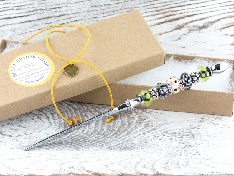 K&ouml;nnte beinhalten: Ein Brief&ouml;ffner mit einem dekorativen, mit Perlen besetzten Griff, pr&auml;sentiert in einer braunen Geschenkbox. Der Griff weist eine Reihe farbenfroher Perlen auf, darunter Schwarz, Wei&szlig; und Gr&uuml;n. Ein gelbes Band mit Herzanh&auml;nger ist an der Box befestigt.