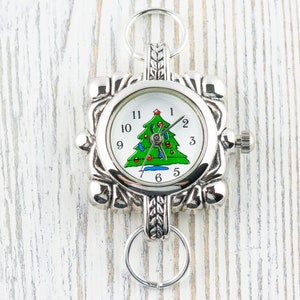 May include: Silver charm watch with a Christmas tree design on the face. The watch has a square face with a silver metal frame and a silver chain.